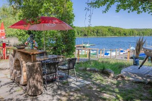 Un espace extérieur aménagé avec une table et des chaises, un parasol rouge et une vue sur un lac où sont amarrés des bateaux.