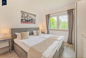 A hotel room with a large bed, two bedside tables, a window with curtains, and a cityscape painting on the wall. - Ferienhaus Zirchow 4 - Modernes, 125 m² Ferienhaus für bis zu 8 Gäste, Garten, Grillmöglichkeiten, (Zirchow)