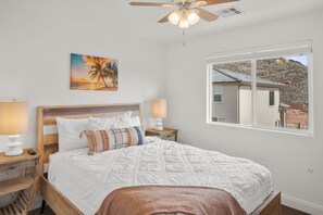A bedroom with a wooden bed, white bedding, a painting of a sunset, and a window with a view of a house and mountains.