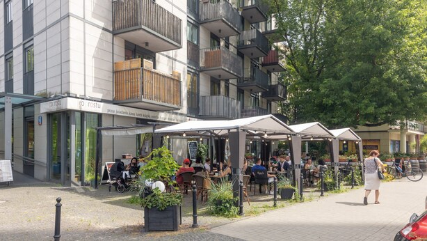 Un área de café al aire libre con mesas y sillas, rodeada por un edificio moderno con balcones.