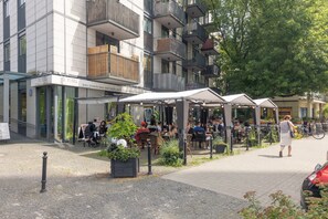 Ein Cafébereich im Freien mit Tischen und Stühlen, umgeben von einem modernen Gebäude mit Balkonen.