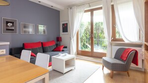 Een moderne woonkamer met een grijze bank, rode kussens, een witte salontafel en een grijze fauteuil. Er hangen ingekaderde foto's aan de muur en is een groot raam met gordijnen.