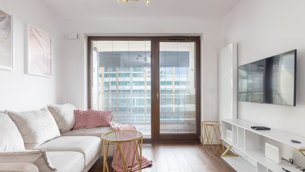 A modern living room with a sofa, a small round table, a TV mounted on the wall, and a sliding glass door leading to an outdoor area. - Burakowska Pastel Elegant Apartment (Warszawa)
