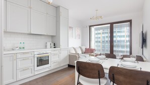 A modern kitchen with white cabinetry, a dining area with a white table and chairs, and a large window with blinds. - Burakowska Pastel Elegant Apartment (Warszawa)