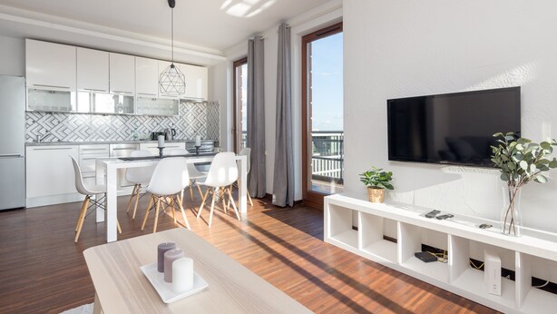 Una cocina moderna con armarios blancos, un comedor con mesa y sillas, un televisor montado en la pared y un balcón con vistas.