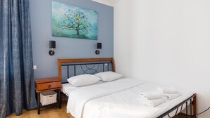 A bedroom with a wooden headboard, a white bedspread, two bedside lamps, a blue curtain, and a painting of a tree on the wall.