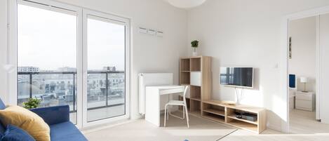 Un apartamento moderno con un amplio ventanal corredero, un escritorio blanco con silla, un televisor y un sofá azul.