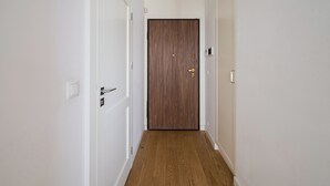 A narrow hallway with white walls and a wooden floor leading to a dark wooden door with a brass handle.