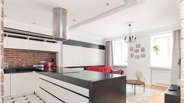 A modern kitchen with white cabinets, a black countertop, and a red chair. There is a brick wall, a window with curtains, and a chandelier. - Old Town Grey Apartment (Warszawa)