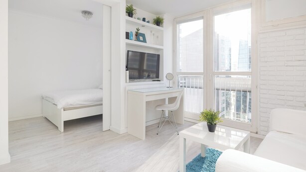 Una habitación moderna con decoración blanca, que incluye una cama, un escritorio con ordenador y estanterías, una mesita con una planta, y vistas a edificios a través de amplios ventanales.