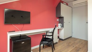 A compact room with a kitchenette, a flat-screen TV mounted on the wall, a black chair, and a white desk.