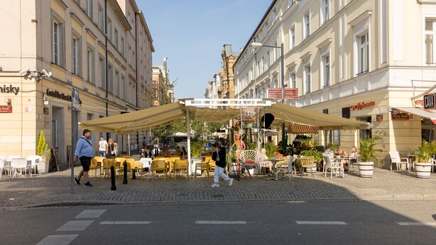 A street with outdoor seating at a cafe, a pedestrian crossing, and buildings with signage.