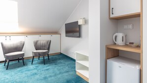 A modern room with two grey upholstered chairs, a white cabinet, a small white refrigerator, a wooden shelf with a kettle, and a flat-screen TV mounted on the wall.