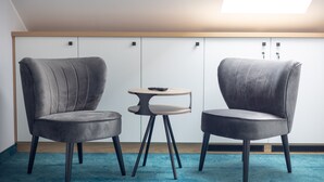 Two grey upholstered chairs with wooden legs, a small round table, and a blue carpet in a room with white walls and a wooden ceiling.