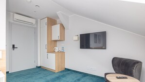 A modern hotel room with a teal carpet, a wooden kitchenette, a grey armchair, and a TV mounted on the wall.