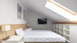 A modern hotel room with a sloped ceiling, a bed with white linens, a bedside table with a lamp, a TV mounted on the wall, and built-in shelving.