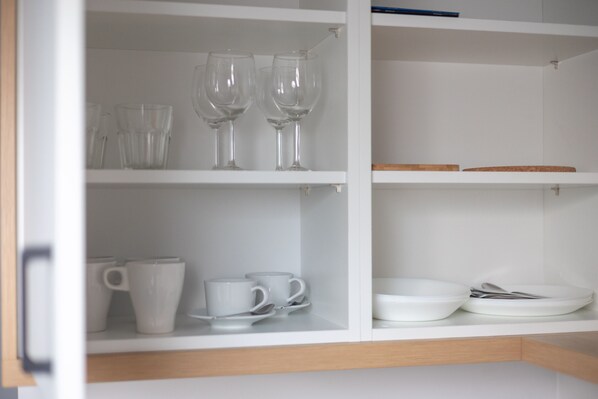 Un armario blanco con estantes que contienen copas de vino transparentes, tazas blancas y platos blancos.