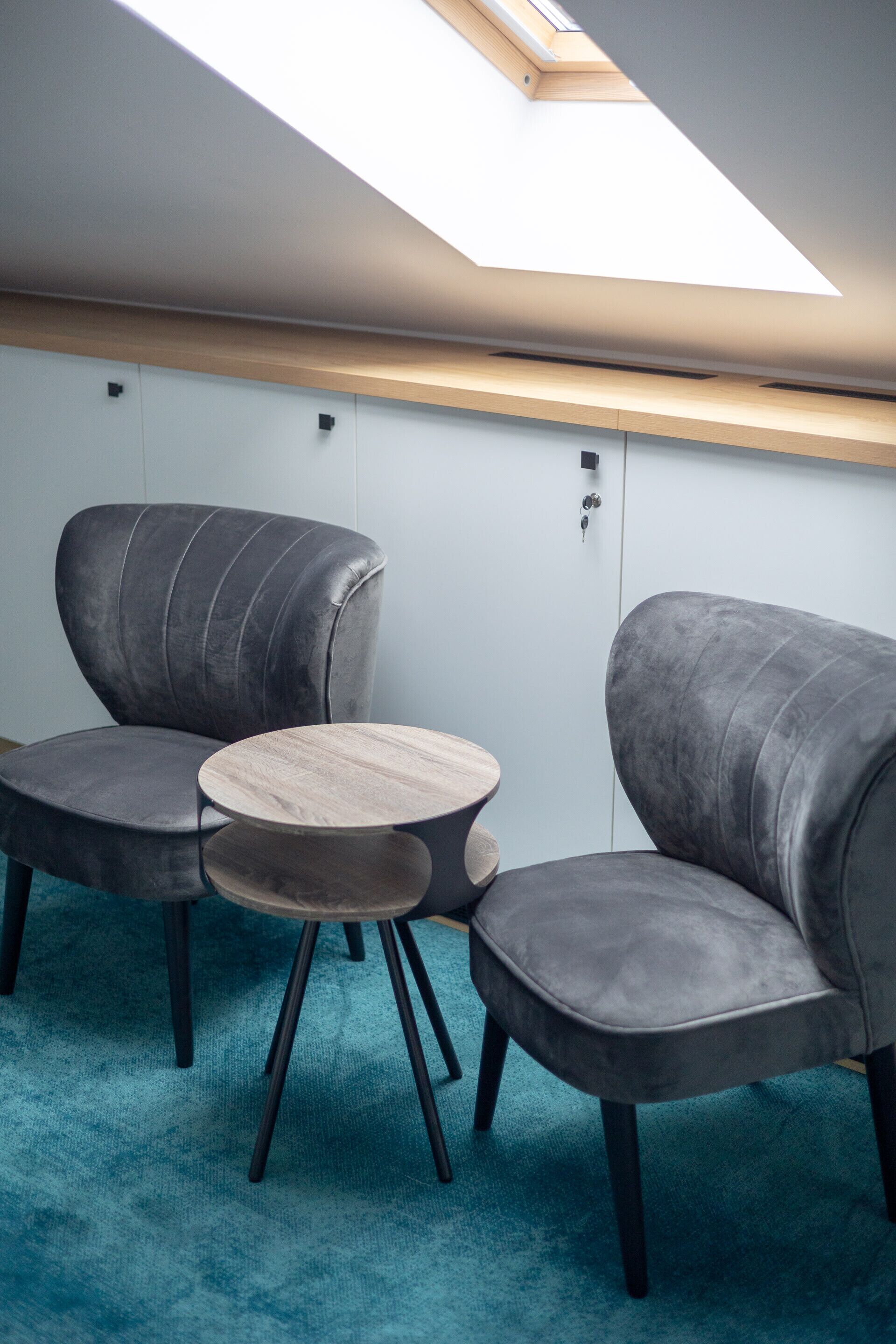 A modern room with two grey upholstered chairs, a round wooden table, and a skylight above.