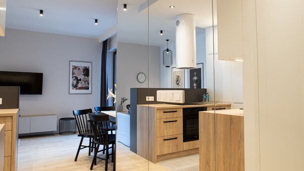 A modern kitchen with wooden cabinets, a dining area with chairs, and a large mirror. - Arkadia Prime Apartament (Warszawa)