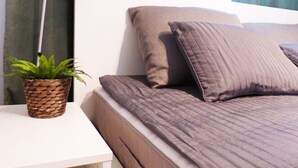 Una cama bien hecha con cabecera, una lámpara de noche y una planta en una mesita blanca.