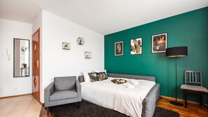 A bedroom with a bed, grey sofa, dark rug, green wall, and framed pictures. - Saska Kępa Malachite Studio (Warszawa)