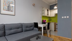 Ein modernes Wohnzimmer mit grauem Sofa, einem kleinen Tisch mit zwei Fernbedienungen, einem gerahmten abstrakten Kunstwerk und einer Küchenzeile mit grünen Fliesen und weißen Schränken.