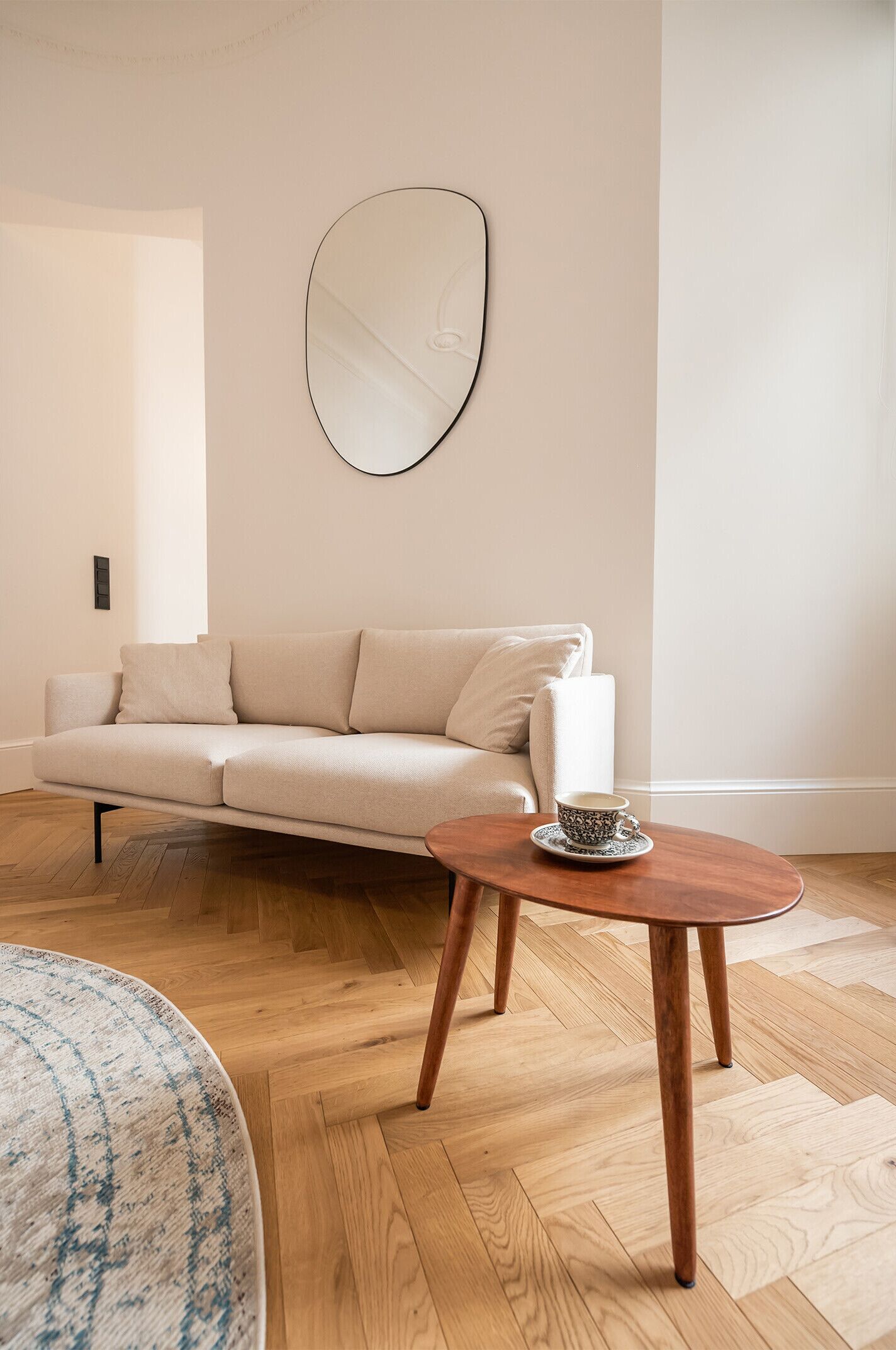A modern living room with a beige sofa, a round wooden coffee table, and a large mirror on the wall.