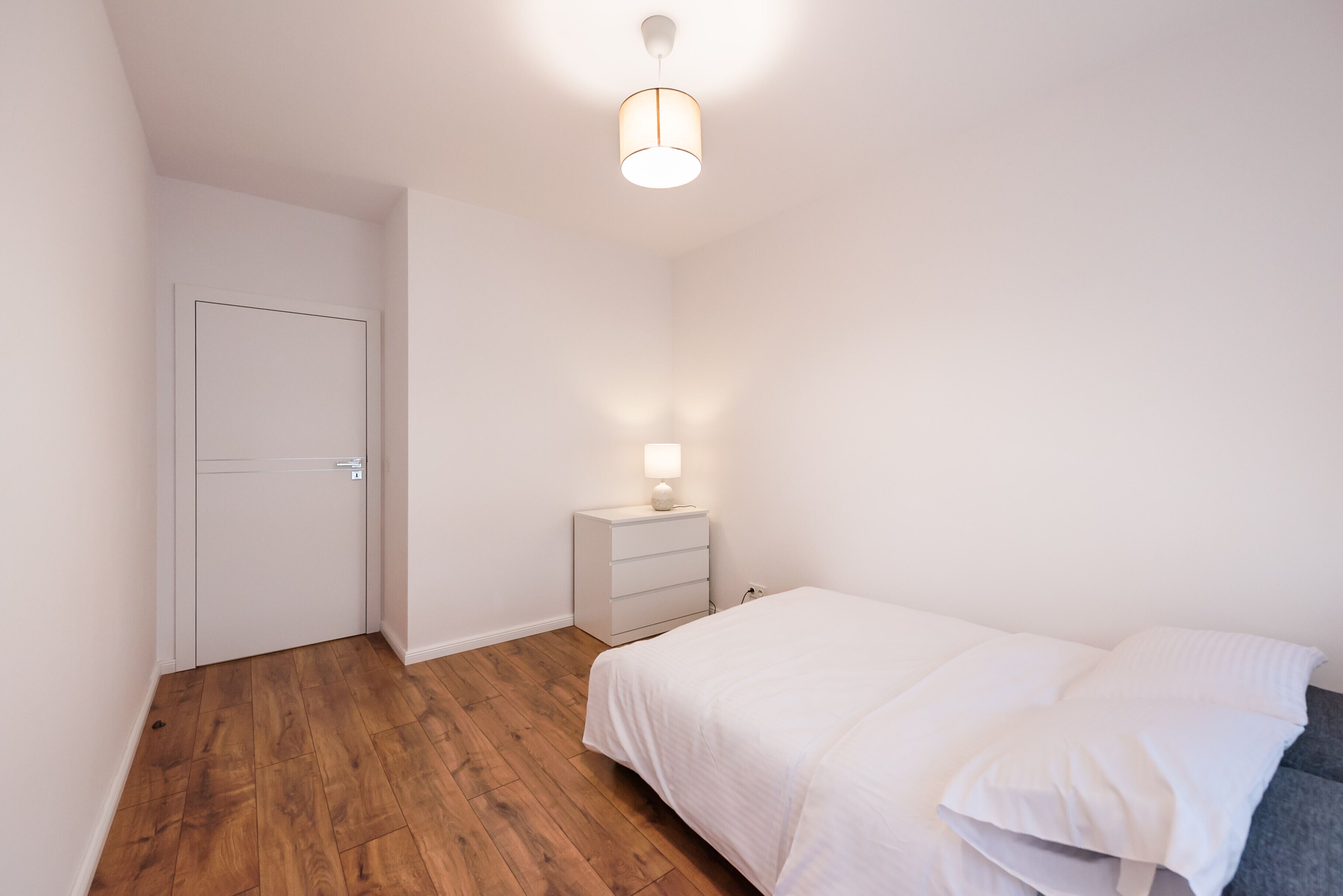 Un dormitorio con piso de madera, una cama con ropa de cama blanca, una mesita de noche con lámpara y una puerta blanca.