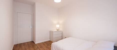 Un dormitorio con piso de madera, una cama con ropa de cama blanca, una mesita de noche con lámpara y una puerta blanca.