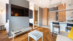 A modern hotel room with a bed, a bench, a small table, a TV, a kitchen area with a bar, and a bathroom visible through an open door. - Nowa Praga Geometric Yellow Apartment (Warszawa)
