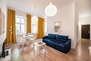 Ein modernes Wohnzimmer mit einem dunkelblauen Sofa, einem weißen Couchtisch und einem Essbereich mit einem weißen Tisch und Stühlen. Zwei Pendelleuchten und ein Fenster mit gelben Vorhängen.