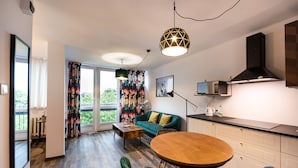 A modern living room with a wooden dining table, green chairs, a sofa with yellow cushions, and a large window with floral curtains. - Ogród Saski Colored Studio (Warszawa)