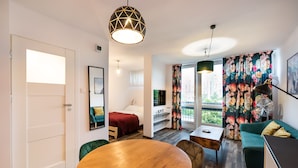 A modern living room with a wooden dining table, a green sofa, a bed with a red blanket, and a balcony with floral curtains. - Ogród Saski Colored Studio (Warszawa)
