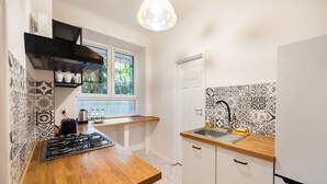 A compact kitchen with a gas stove, wooden countertop, black hanging shelf, patterned backsplash, and a window with greenery outside. - Metro Plac Wilsona Etno Apartament (Warszawa)