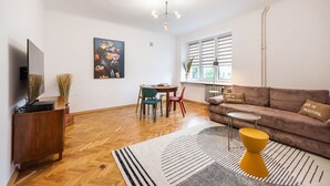 A modern living room with a flat-screen TV, a sofa with decorative pillows, a dining area with a table and chairs, and a large window with blinds. - Metro Plac Wilsona Etno Apartament (Warszawa)