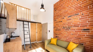 A room with a loft bed, a wooden desk, a green sofa with yellow cushions, a brick wall, and a hanging light fixture. - National Stadium Cosy Studio #3 (Warszawa)