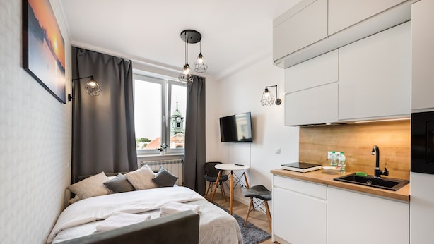 A compact hotel room with a bed, a small dining table, a kitchenette, and a TV mounted on the wall.