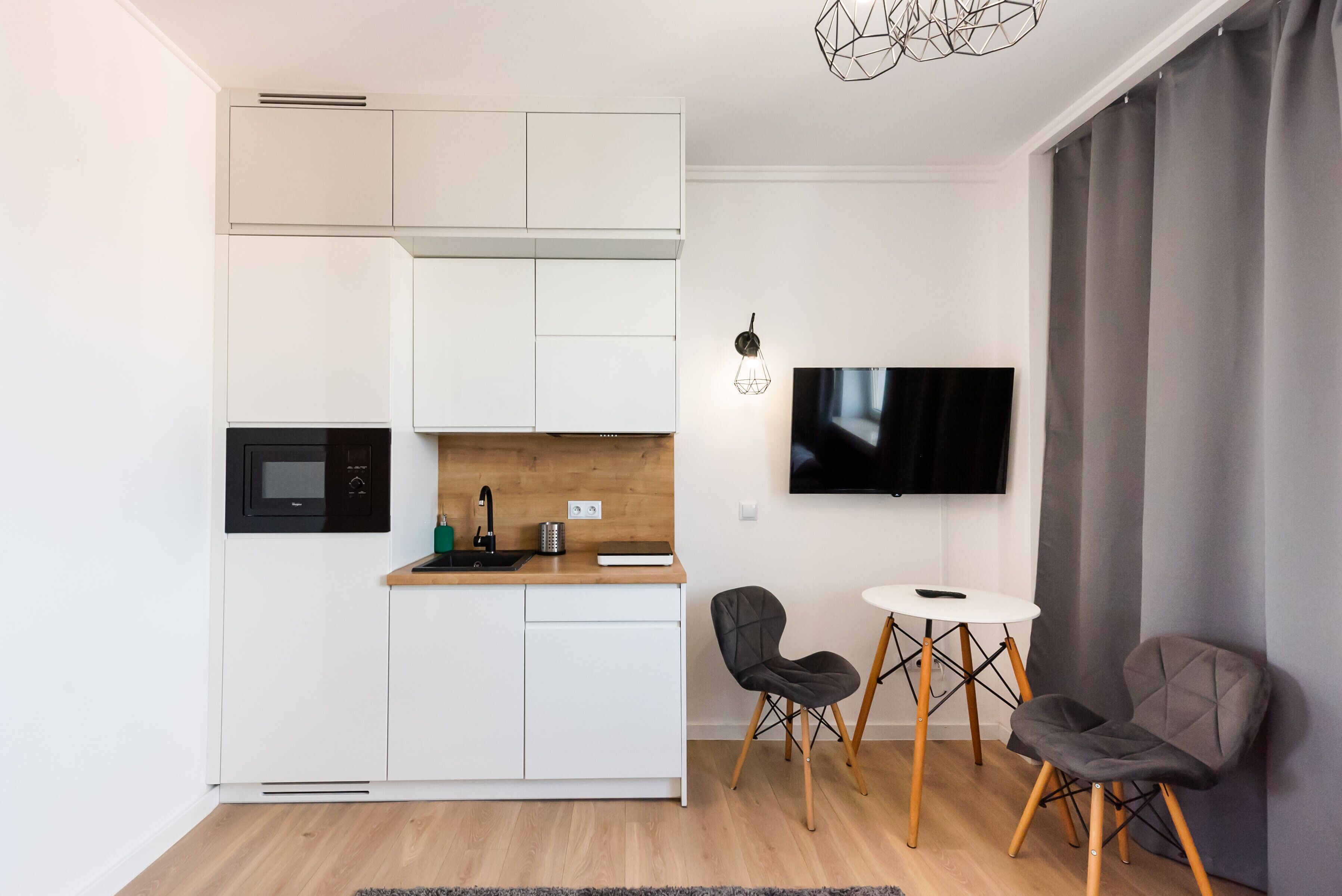 Une cuisine compacte avec des meubles blancs, un four à micro-ondes et un plan de travail en bois. On y trouve un coin repas avec deux chaises et une petite table. Un téléviseur à écran plat est fixé au mur.