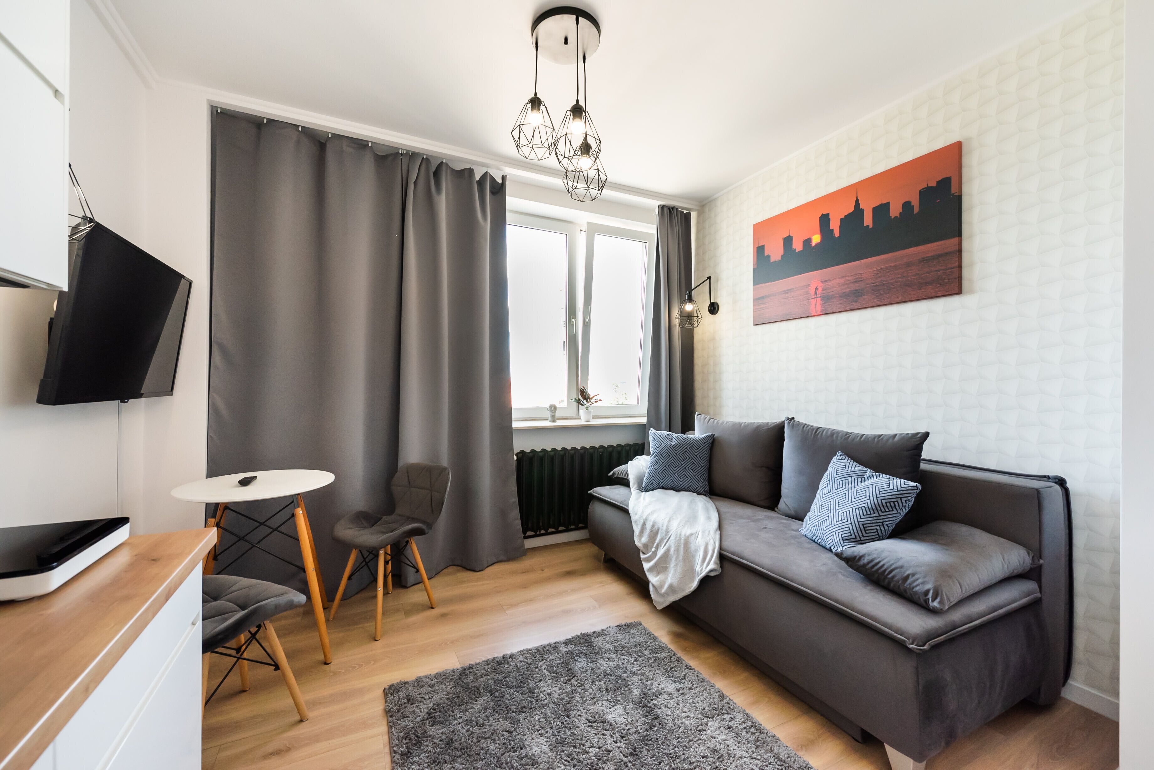 Un salon moderne avec un canapé gris, une petite table ronde, un fauteuil gris, un tapis gris et un tableau représentant un paysage urbain accroché au mur.