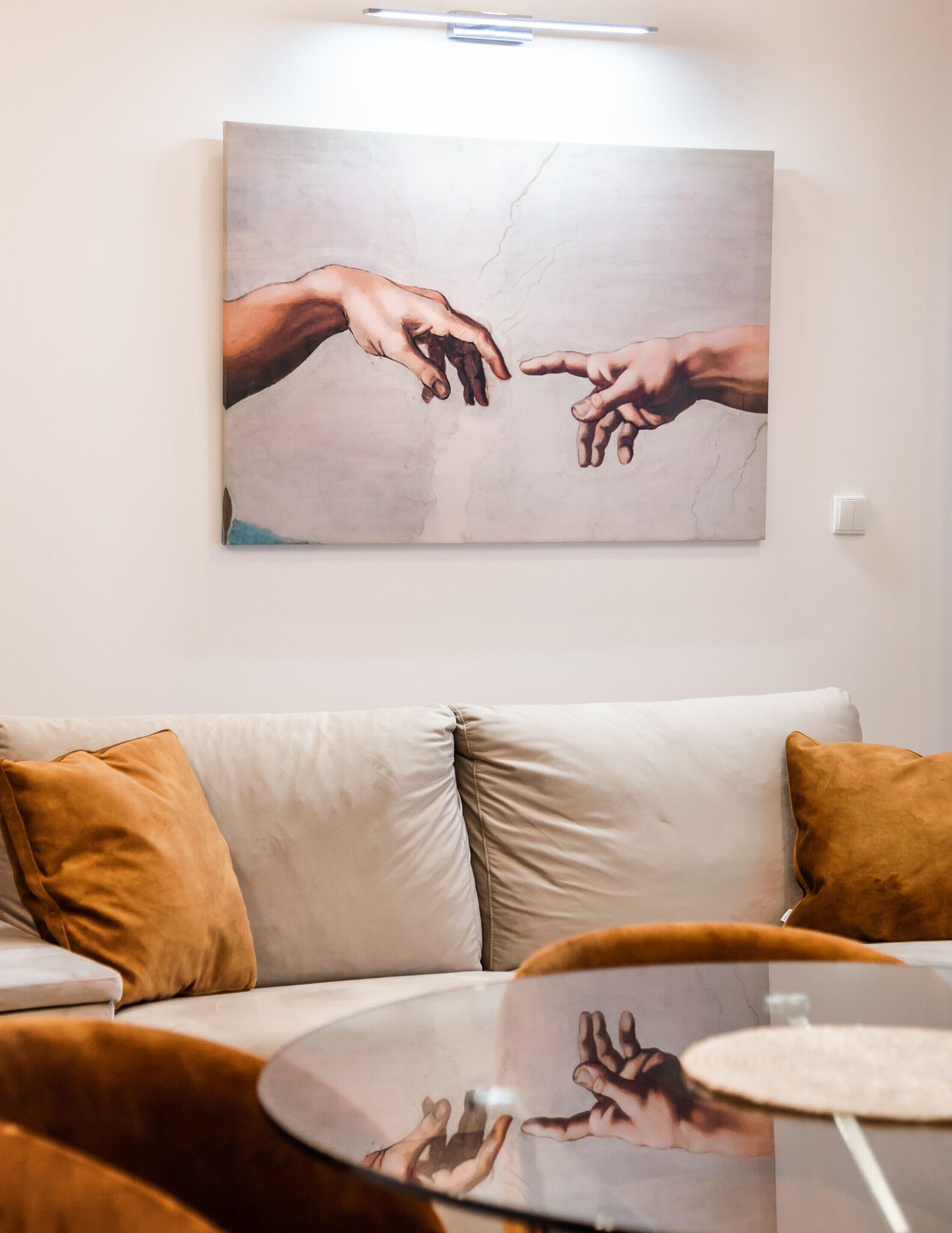 Una sala de estar con un sofá color beige, dos cojines marrones y una mesa de centro de vidrio. En la pared, un cuadro grande de manos extendiéndose la una hacia la otra.