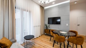 A modern living room with a sofa, a glass table, and chairs, a TV mounted on the wall, and a large window with curtains. - Złota Sophisticated Apartment (Warszawa)