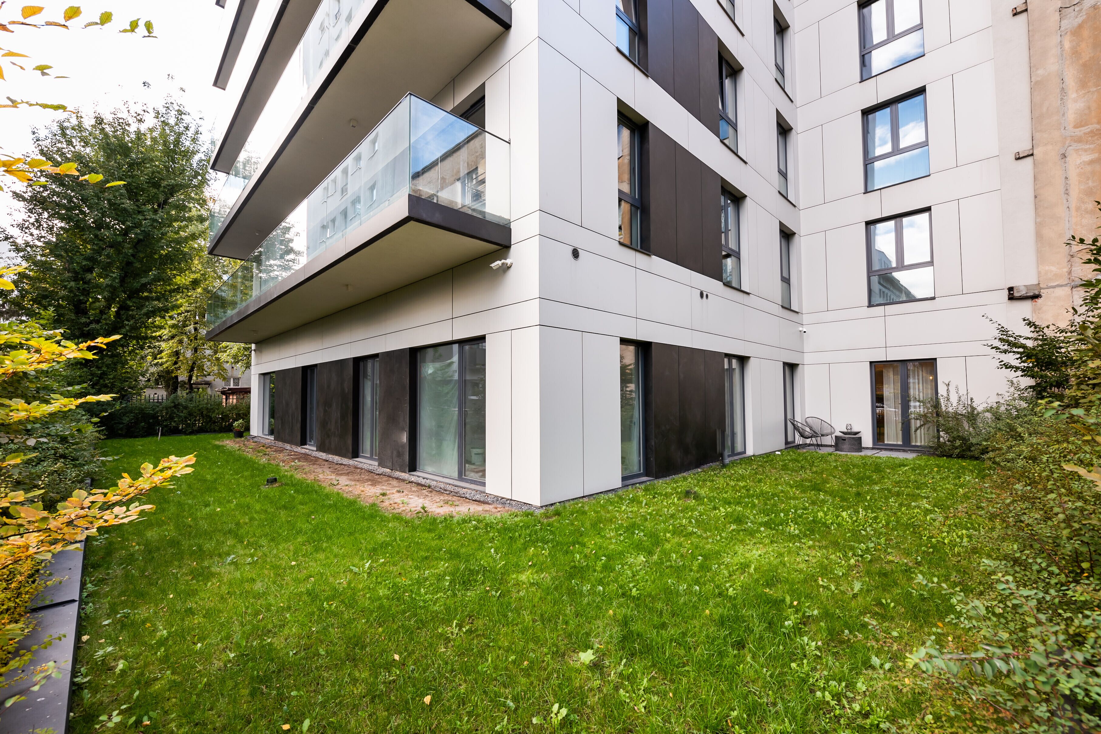 Un edificio moderno de varios pisos con techo plano, ventanas amplias y un pequeño jardín con césped y árboles.