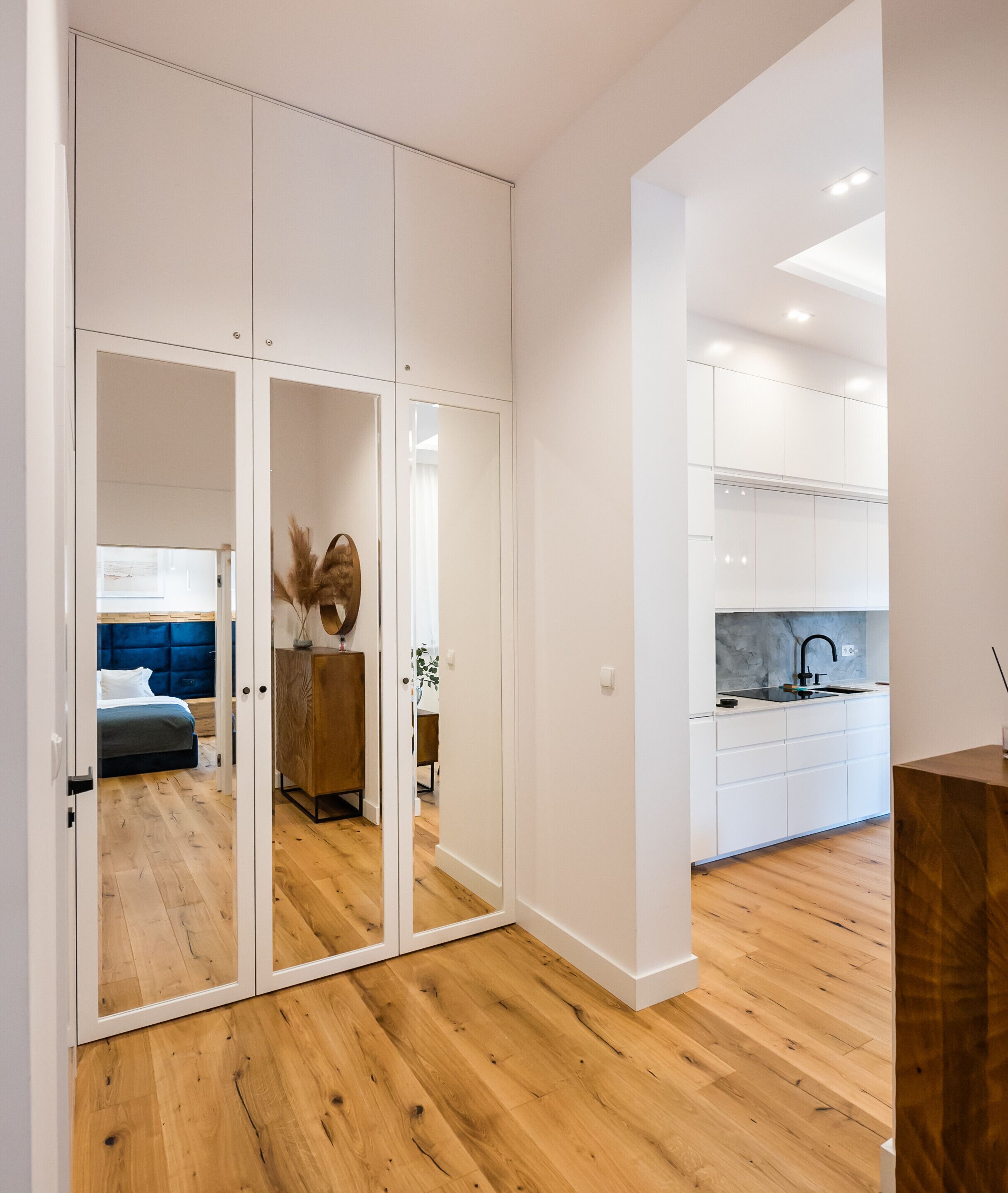 A modern apartment with a wooden floor, a bedroom with a blue headboard, a kitchen with white cabinets, and a mirrored wardrobe.
