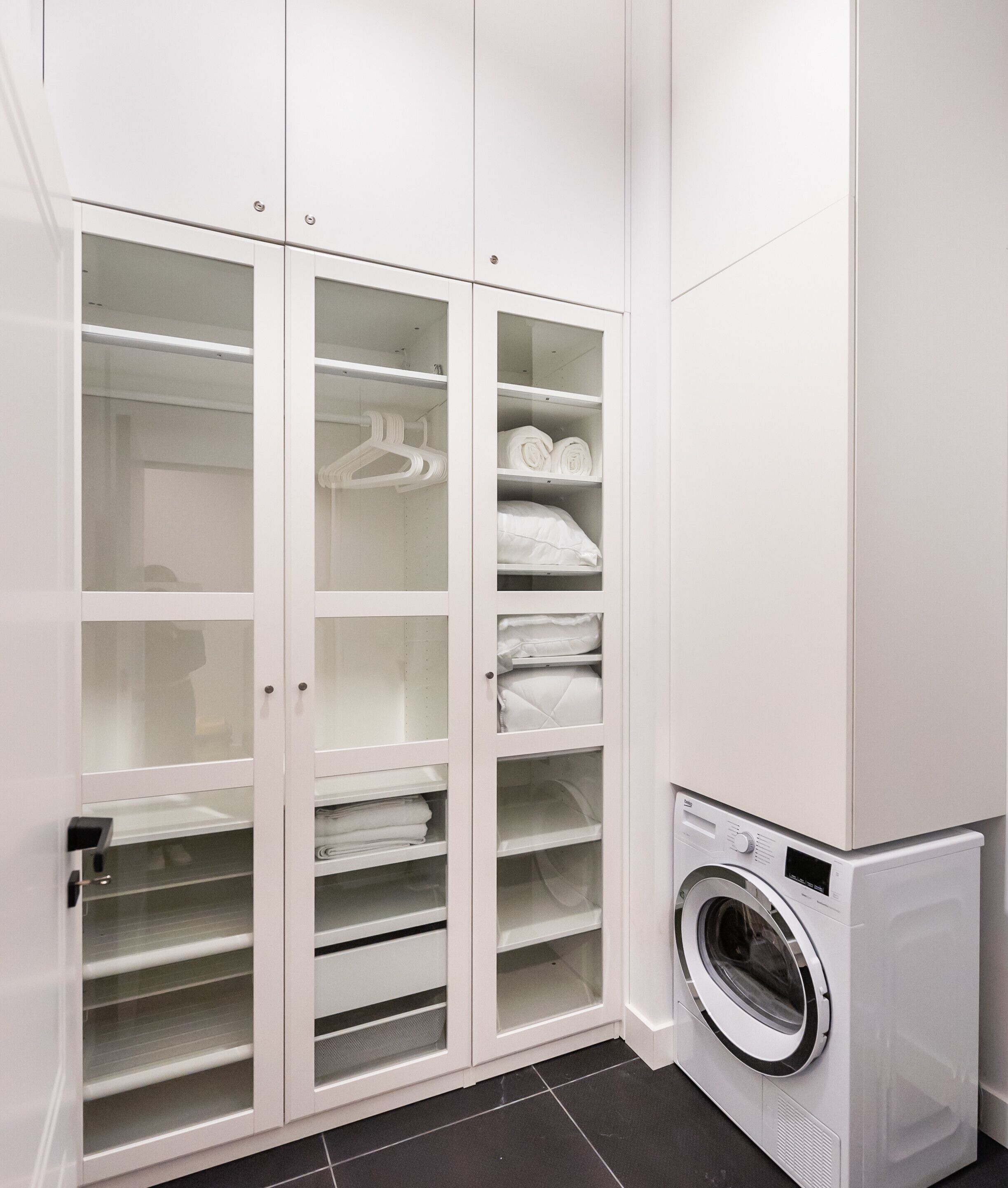 A laundry room with a washing machine and a white storage closet with multiple shelves and hanging space.