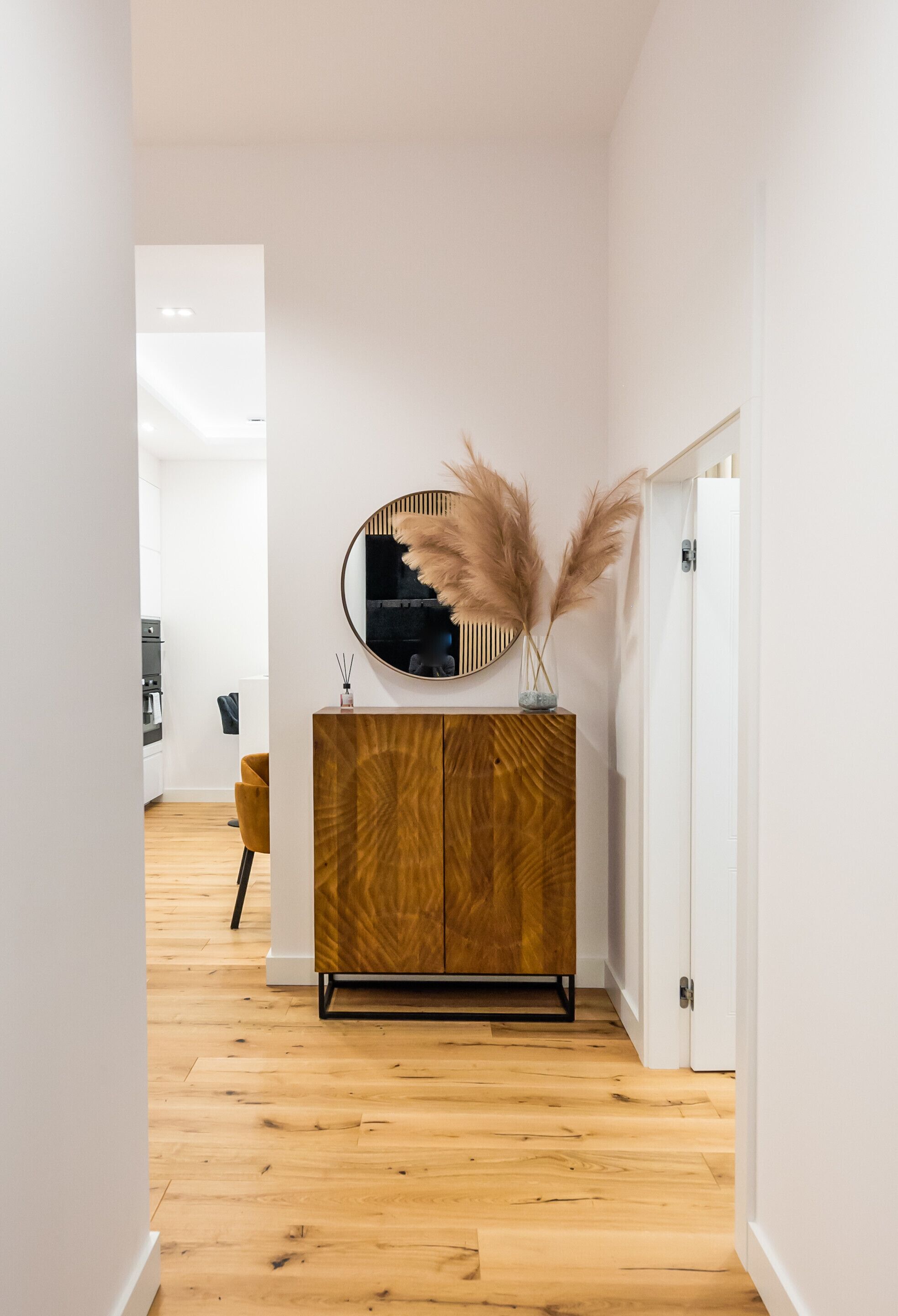 A hallway with a wooden floor, a white door, and a wooden cabinet with a mirror and pampas grass decoration.