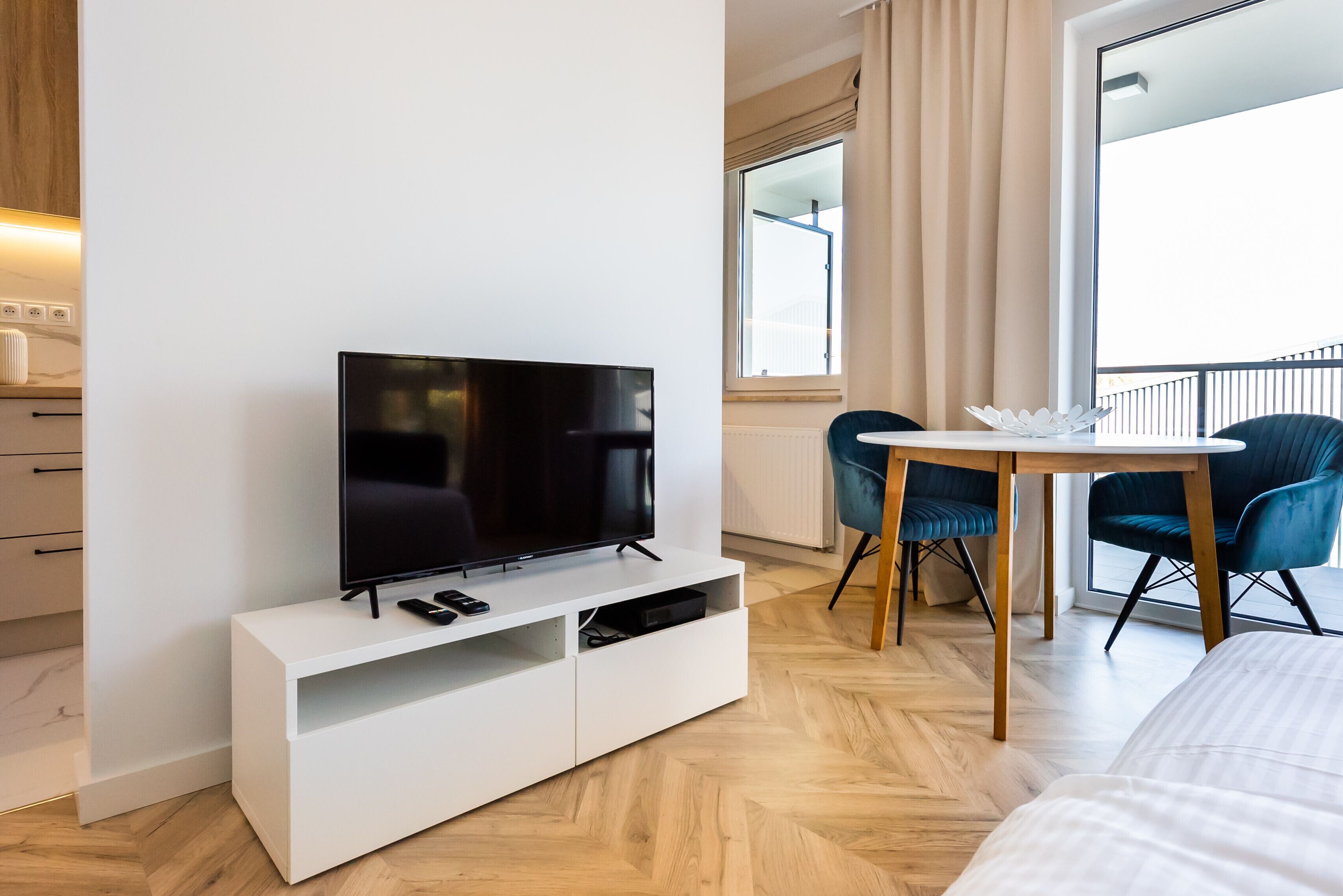 Habitació d'hotel moderna amb televisió de pantalla plana sobre una consola blanca, zona de menjador amb taula i cadires, i una finestra gran amb vistes a l'exterior.