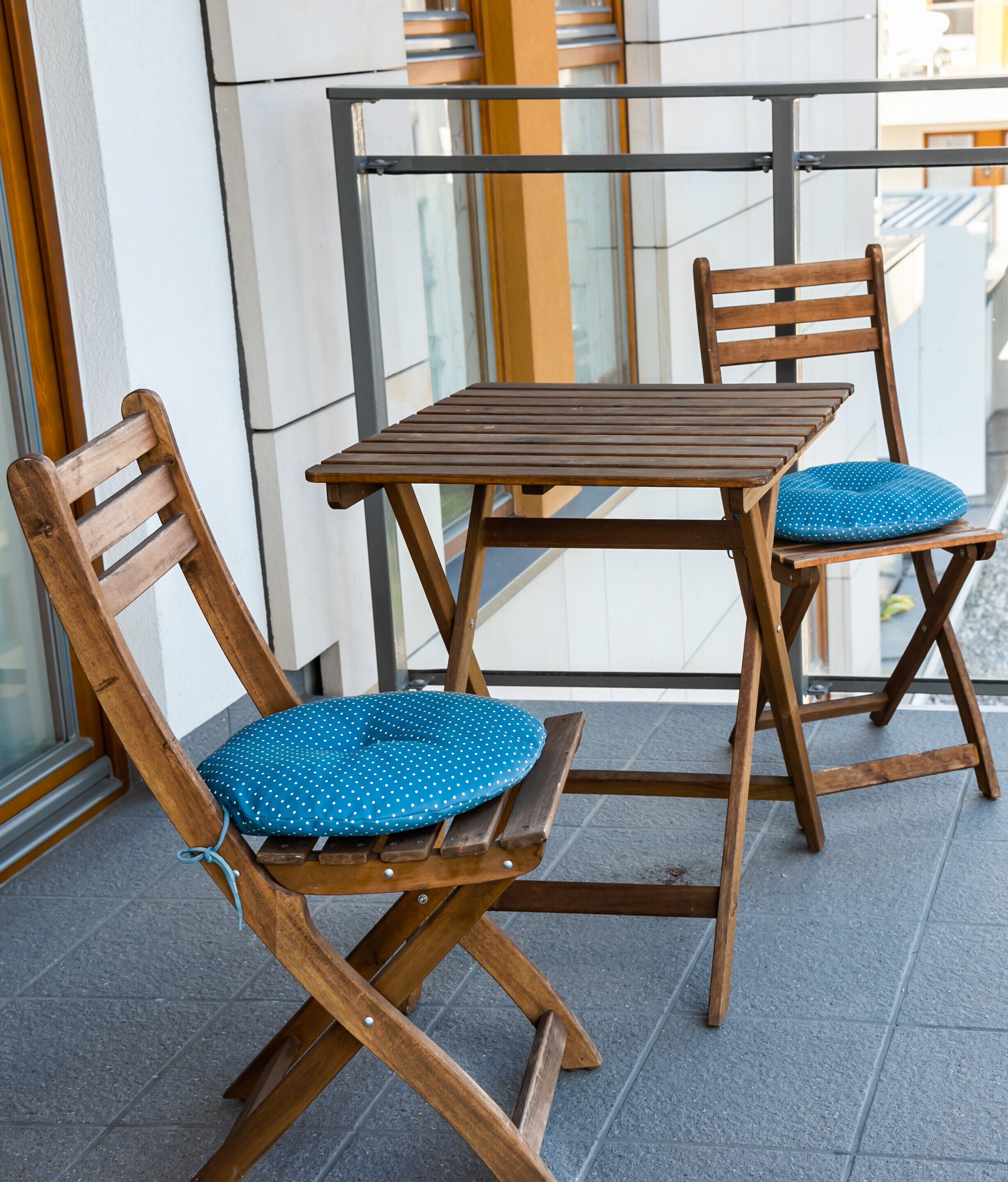 A wooden table and two chairs with blue polka dot cushions on a balcony with a glass railing.