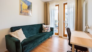 A living room with a green sofa, a wooden table, and a chair. There is a painting on the wall and a view of the outside through a glass door. - Służewiec Comfort Zone Apartment (Warszawa)