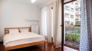 A bedroom with a wooden bed, white bedding, a radiator, and a view of an apartment building through a glass door. - Malborska Lovely Apartment (Warszawa)
