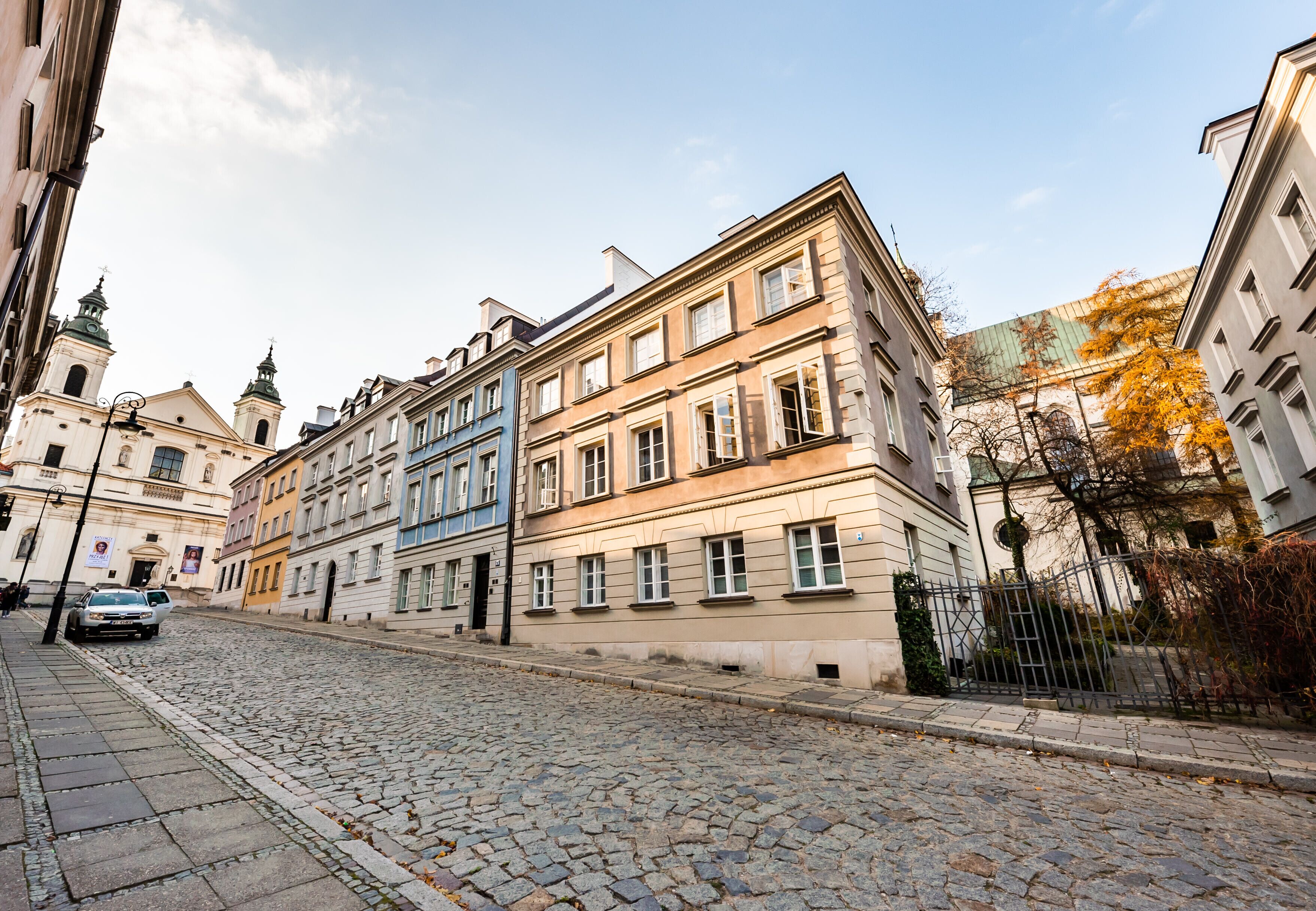 Eine gepflasterte Straße gesäumt von historischen Gebäuden, darunter eine Kirche mit einem Glockenturm und ein mehrstöckiges Wohngebäude mit großen Fenstern.